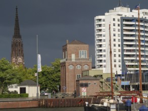 A cityscape with buildings, a church tower and ships in the harbour under a cloudy sky,