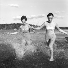 1950s summer. Two young women runs on the shallow part of the beach and the water splashes. Sweden