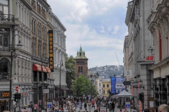 Busy shopping street with several shops, in the background a hilly panorama and cloudy sky, oslo,