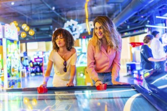 Two female friends concentrating while playing air hockey in a vibrant arcade, enjoying their