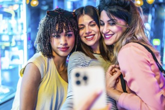 Three cheerful female friends are taking a selfie with a smartphone in a vibrant, illuminated urban