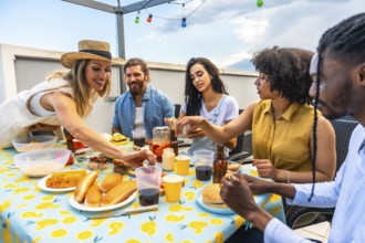 Happy multi ethnic friends having lunch together on urban rooftop, eating hot dogs, burgers, and