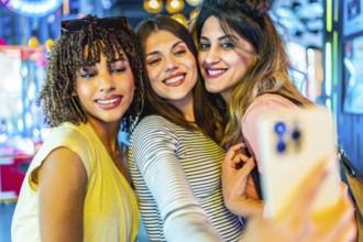 Smiling friends capturing a joyful selfie with a smartphone in a vibrant amusement arcade filled