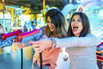 Two women laughing and enjoying their free time while playing arcade games with colorful toy guns,