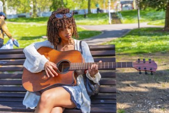 Musician concentrating while playing acoustic guitar sitting on a bench in a green city park during