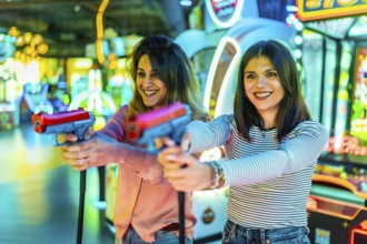 Two happy women enjoying their free time playing together in an amusement arcade holding toy guns
