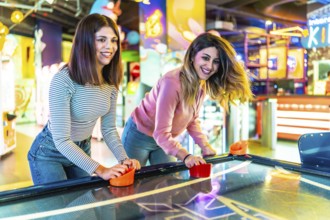 Two female friends enjoying their free time playing air hockey in a colorful and modern amusement