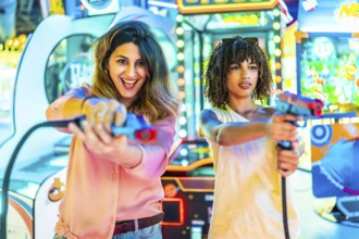 Two women enjoying their free time playing in an arcade with toy guns, laughing and having fun