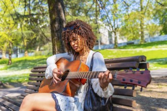 Curly haired woman sitting on a bench playing an acoustic guitar in a green city park during a