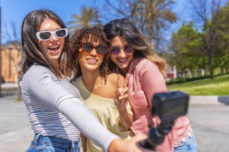 Three cheerful young women making peace sign and taking selfie with action camera during summer