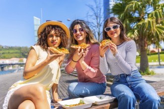 Three women enjoying pizza slices while sitting on a bench in a sunny city park, sharing laughter