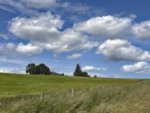 Wide green meadow under a sky with imposing clouds and a rural building in the distance,