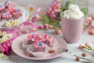 Pink petit fours with flower decoration on a plate next to a cup of coffee with cream