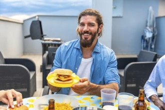 Happy man holding a burger during a rooftop barbecue party with friends, enjoying good food and