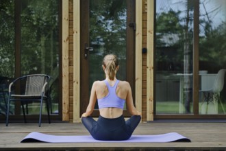Back view of a woman sits in lotus position on a yoga mat placed on a wooden terrace of tiny house