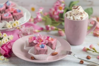 Pink petit fours with flower decoration on a plate next to a cup of coffee with cream