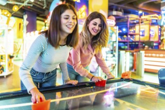 Two young women enjoying their free time playing air hockey in a colorful and modern amusement
