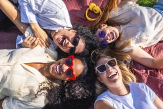 Four cheerful young women wearing sunglasses are lying together on a blanket in a park, happily