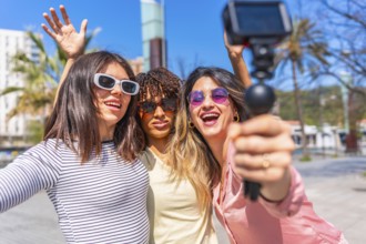 Three cheerful women holding action camera and waving at camera while vlogging their summer