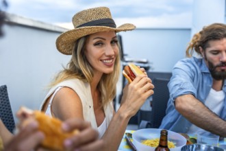 Happy woman eating hot dog at a rooftop barbecue party with friends, enjoying good food and company