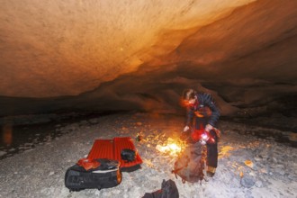 A person kneels by a campfire in a glacier cave, surrounded by darkness and warm light
