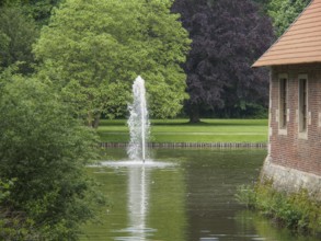 Fountain in a green park next to a brick building, with large trees and calm water, havixbeck,