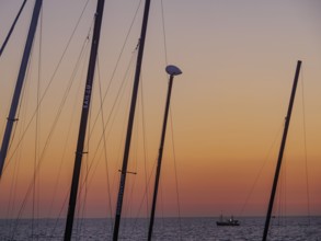 Masts in the foreground and a small boat in the sea at sunset, the sky is bathed in orange tones,