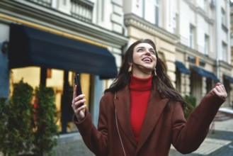 Excited trendy woman celebrating good news holding phone in the evening street