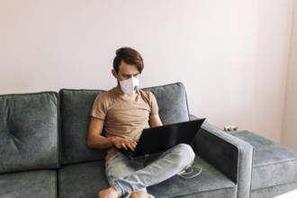 Man in a mask actively using a laptop while sitting on a sofa