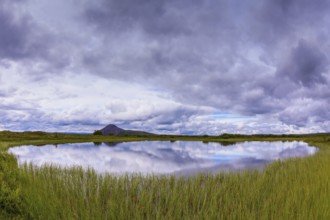 Small lake surrounded by green meadow. In the smooth water of cold lake reflects cloudy sky. Summer