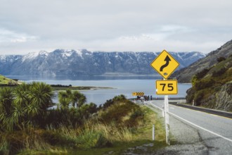 Road with danger signs leads along the lake in front of snow-covered mountains, Wanaka, New Zealand