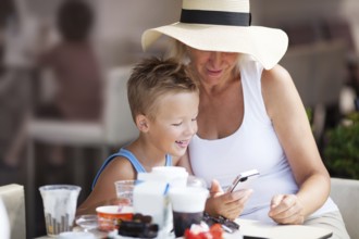 Smiling Mature Woman and Young Boy On Vacation Using Cell Phone Together on Outdoor Restaurant