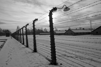 A black and white shot of the outer barbed-wire fence and huts under snow in winter at the