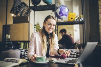 Beautiful young caucasian woman drinking coffee and typing on a keyboard inside a cafe at a wooden