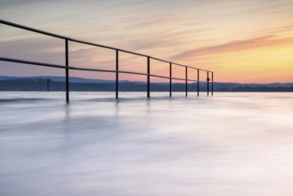 A railing in calm water, surrounded by a pastel-coloured sunrise, lido, Litzelstetten, Lake