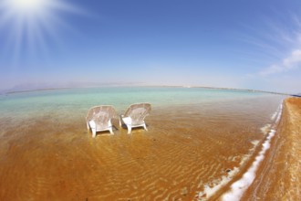 Two white beach chairs stood side by side in the clear water. The sun shines on the beach