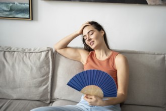 Satisfied woman smiling, blowing with fan, sitting on sofa in home studio. Happy pleased girl