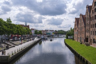 City view with waterway and historic brick buildings, flanked by green areas and clouds, Lübeck,