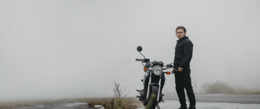 A young man stands by a motorcycle on a foggy road, looking ahead