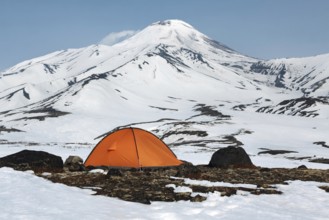 Orange camping (tourist) tent stands on meadow surrounded by snow at foot of beautiful active