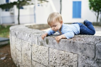 Kid looks behind the stone fence in the yard, lying on it.