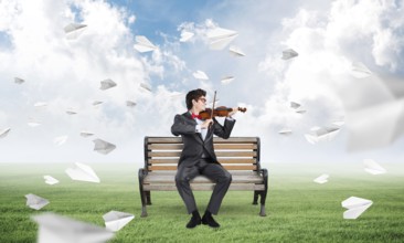 Young businessman plays the violin sitting on a bench, around flying paper planes. Creativity in