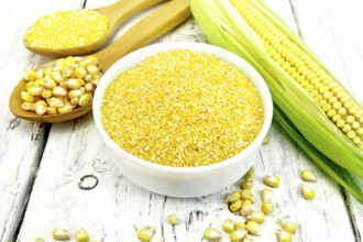 Corn grits in a white bowl and spoon, cobs and grains on the background of wooden boards