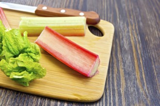 Rhubarb stalks with green leaves and a knife on a small planch on a wooden boards background