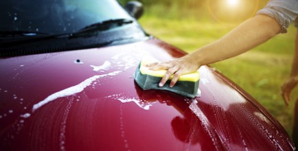 Hand cleaning a red car with sponge under sunlight
