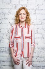 A beautiful young woman student with red curly hair and freckles is leaning against a brick wall of