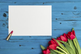 Red tulips, lipstick and a white sheet of paper for your greetings on the background of blue wooden