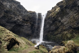 The groom circles the bride in his arms near the waterfall, it is snowing. Destination Iceland