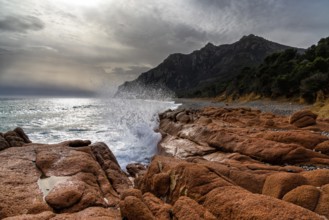Wild rugged coast in Sardinia with waves crashing onto red granite boulders in the foreground
