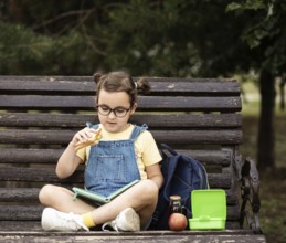 Cute little schoolgirl eating from lunch box outdoor sbtting on a bench. Food for kids